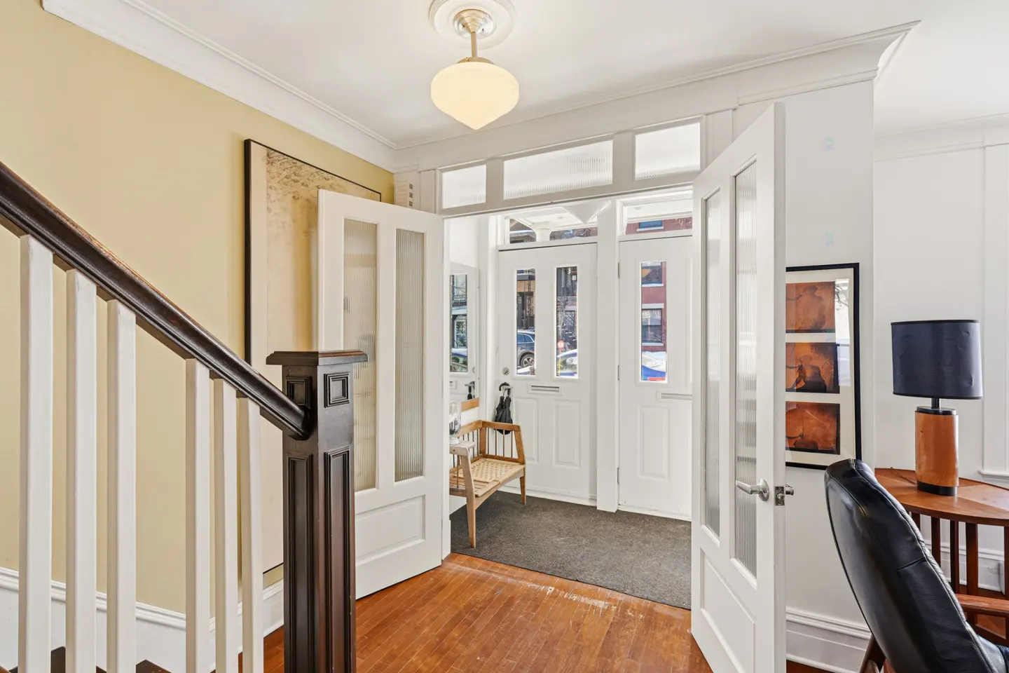 Foyer with open white doors, wood floors, and a staircase with white railings. A bench sits near the front door. A lamp and chair are visible.