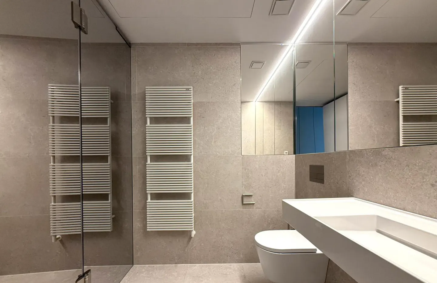 Modern bathroom with beige tile walls, white toilet and sink, and two white towel warmers. A large mirror reflects the room.