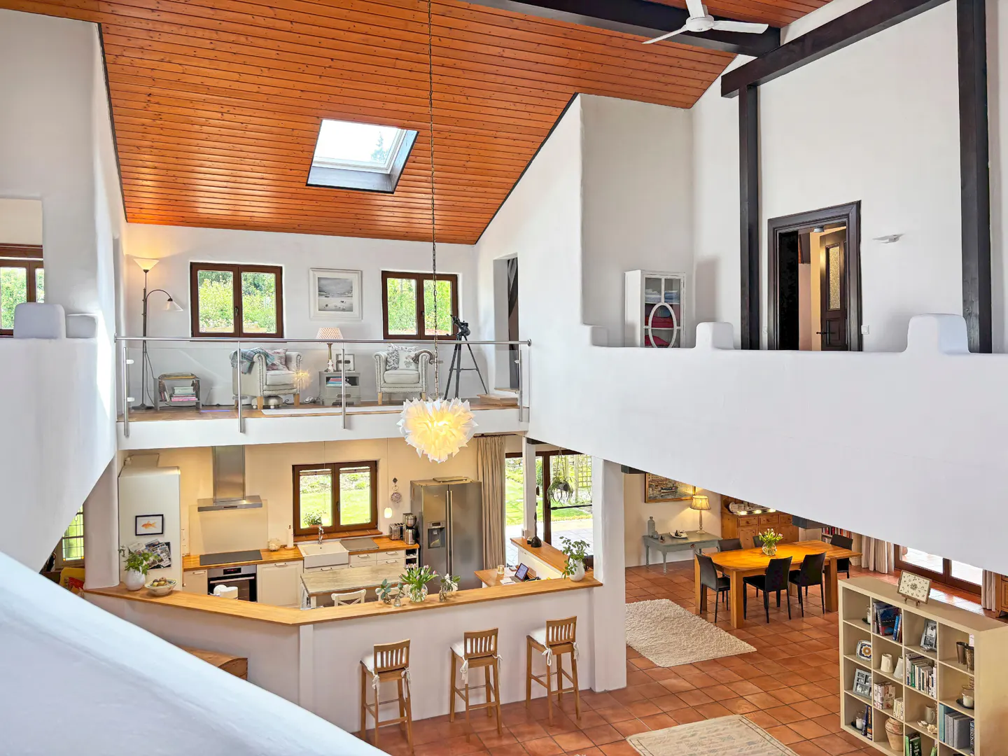 Open-concept home interior with a wood ceiling, white walls, and terracotta tile floors. A kitchen, dining area, and loft are visible.