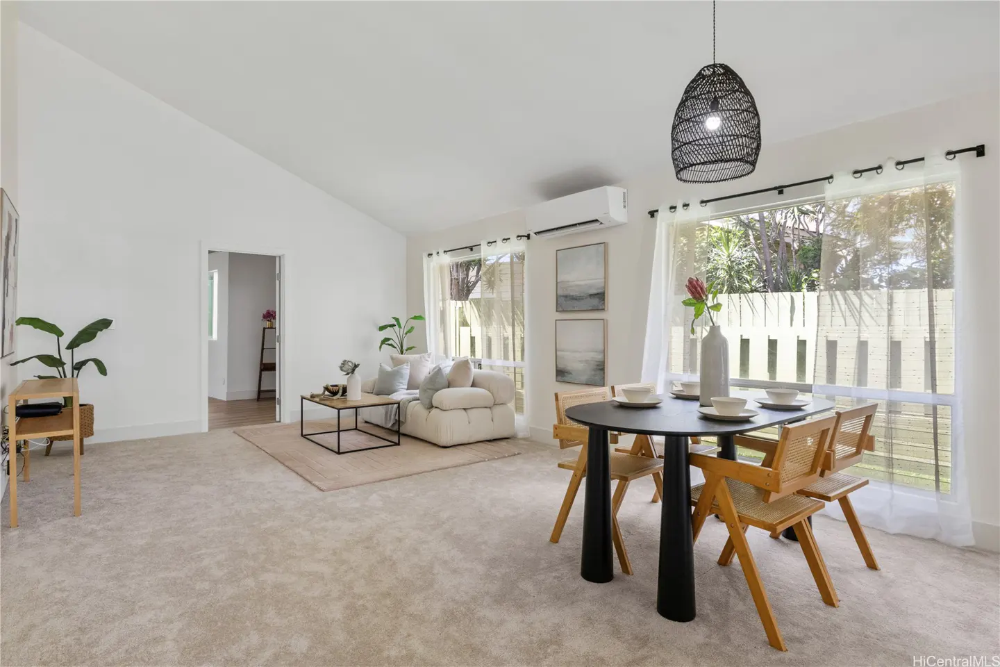 Bright, open-concept living and dining area with beige carpet, white walls, and natural light. A black dining table with wooden chairs sits near a window.
