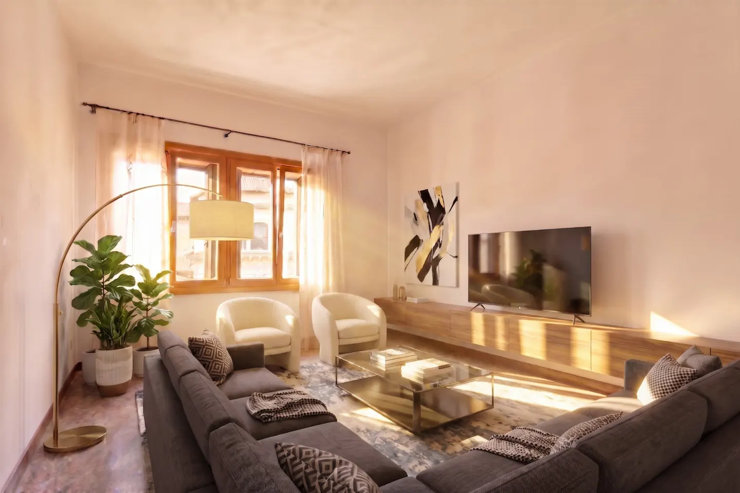 Modern living room with a grey sectional, two cream armchairs, a glass coffee table, and a wall-mounted TV in warm light.