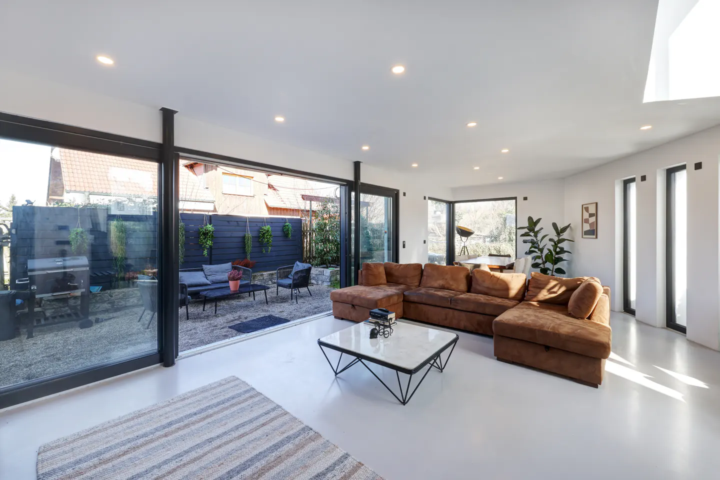 Bright living room with white walls, brown sectional sofa, and a modern coffee table. Sliding glass doors open to a patio with outdoor seating.