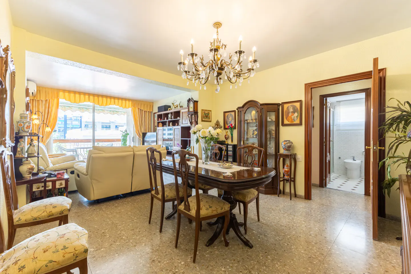 A bright living and dining area with a chandelier, wood furniture, and a view to a bathroom.