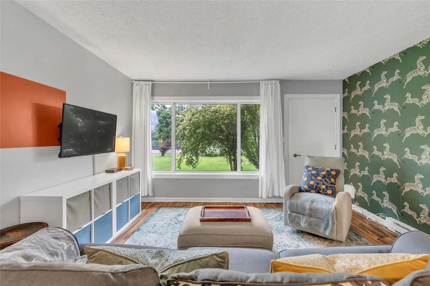Living room with a TV, white storage unit, beige ottoman, recliner, and a green wall with a white zebra pattern.