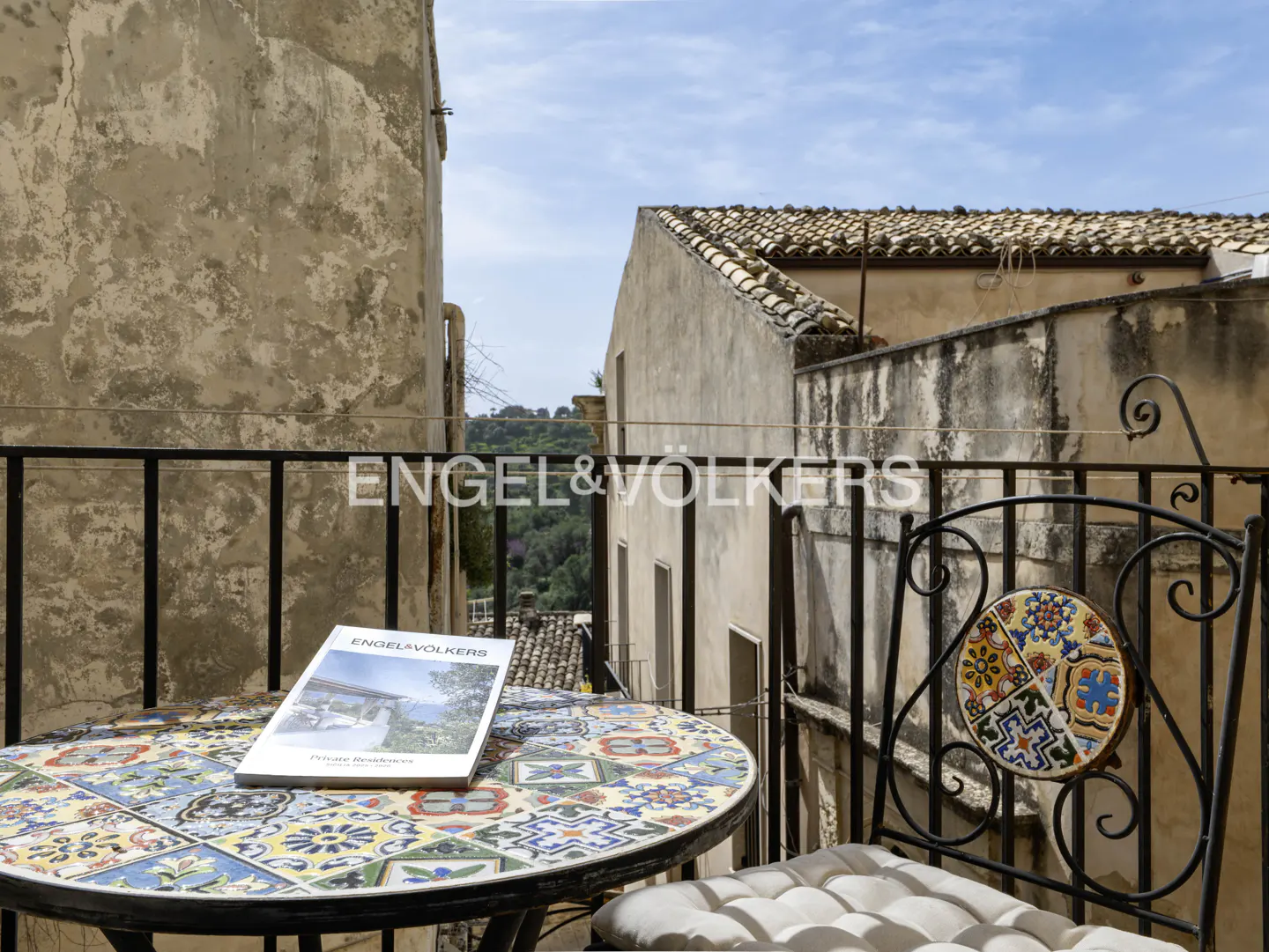 Balcony view with a colorful tiled table and chair. An Engel & Völkers brochure sits on the table. Old buildings and greenery are in the background.