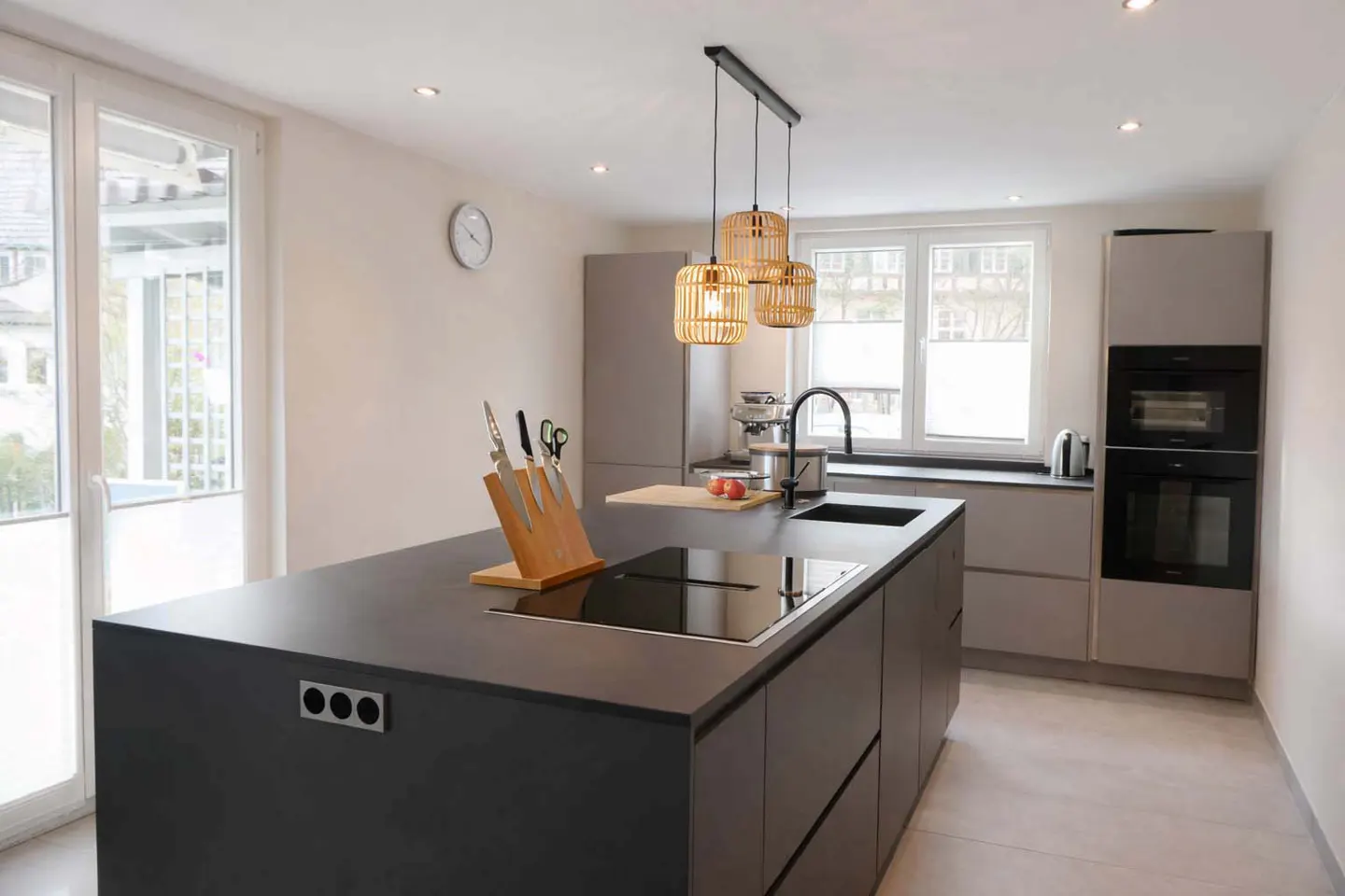Modern kitchen with a black island, sink, cooktop, and bamboo pendant lights. Gray cabinets and stainless steel appliances in the background.
