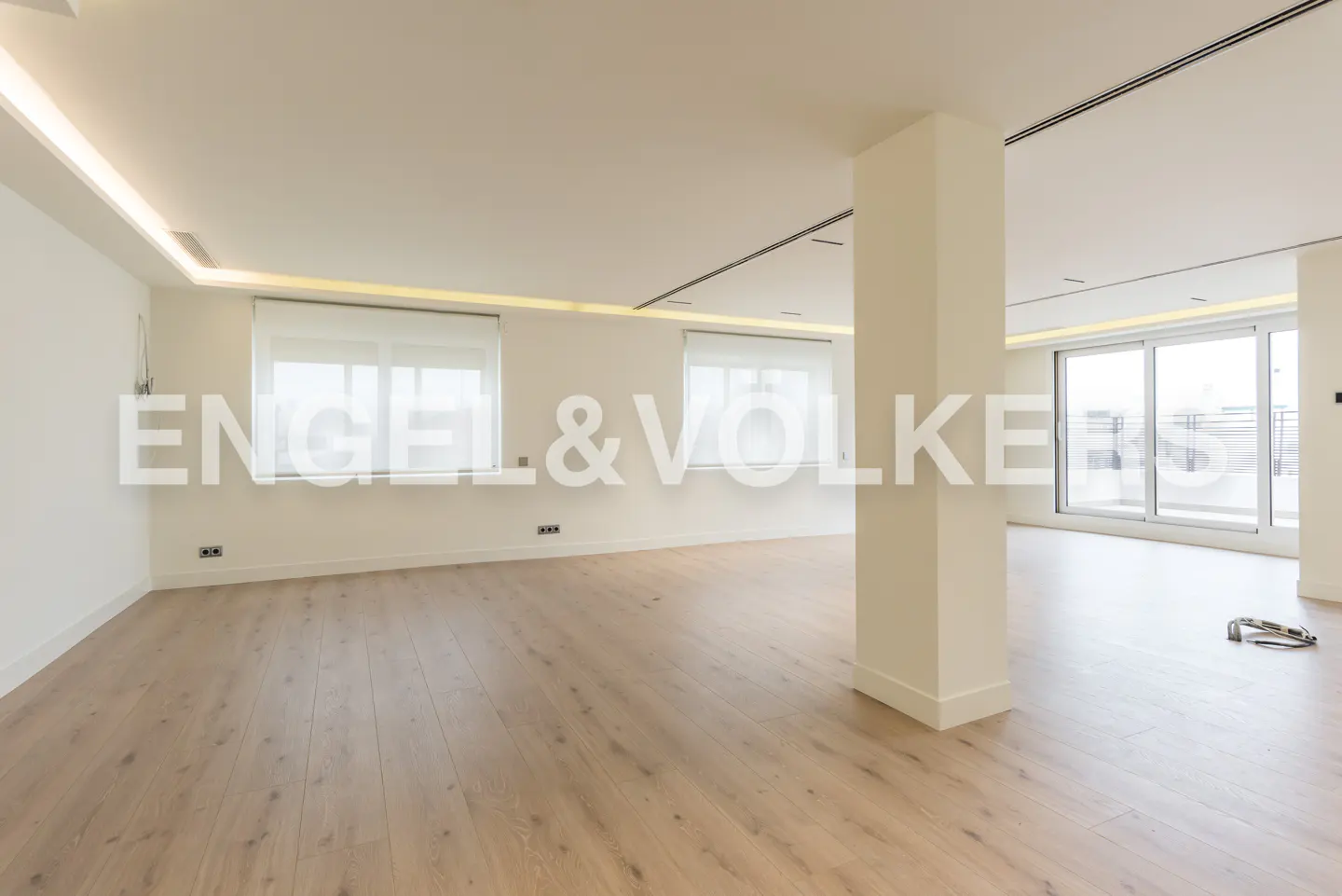 An empty, bright room with light wood floors, white walls, and large windows. A support column stands near sliding glass doors to a balcony. "Engel & Volkers" logo overlay.