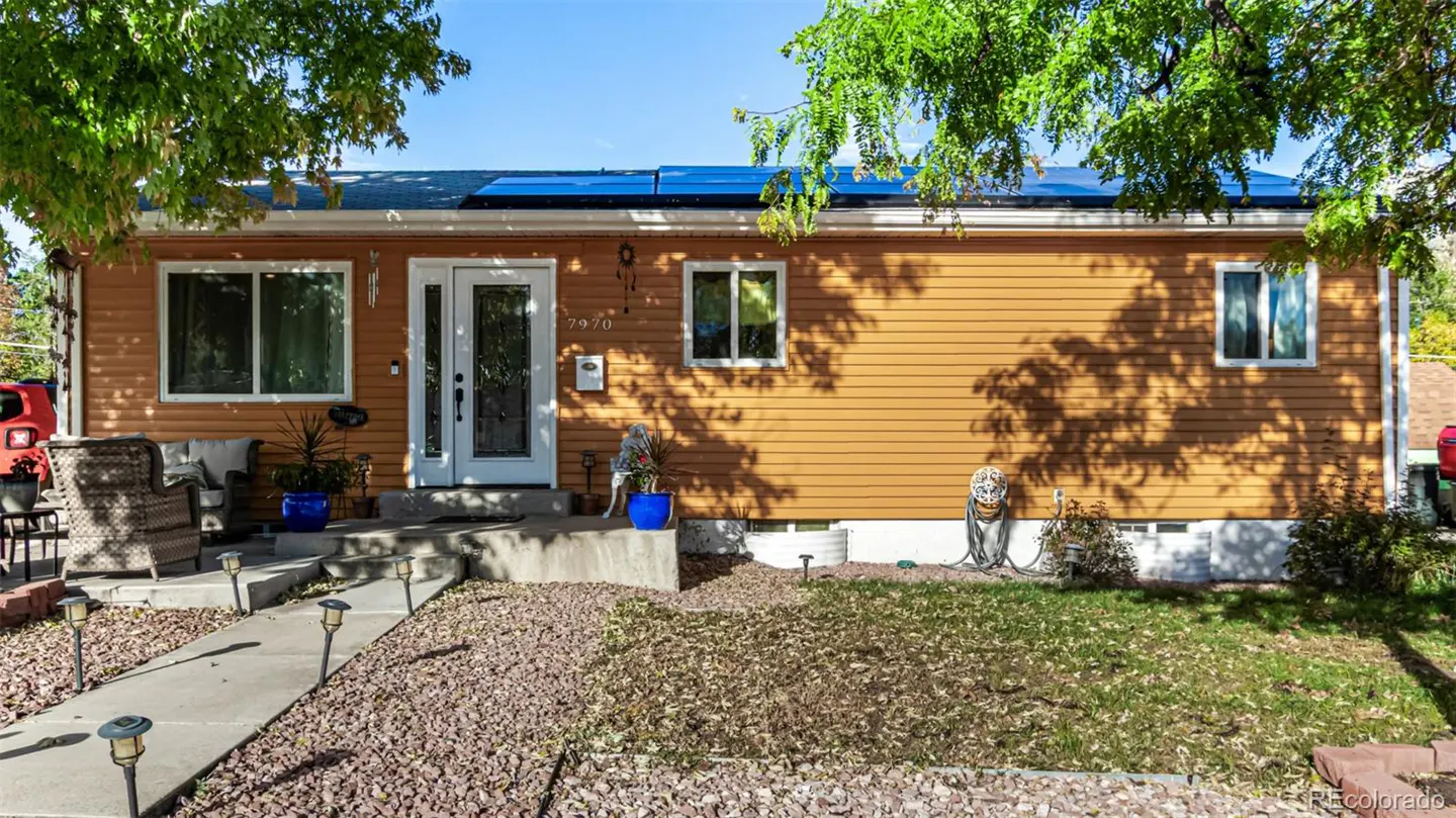 A single-story house with orange siding, white trim, and solar panels on the roof. A stone path leads to the front door.