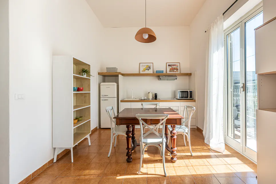 Bright kitchen with a dark wood table, white chairs, and a retro fridge. Sunlight streams in from the balcony door.