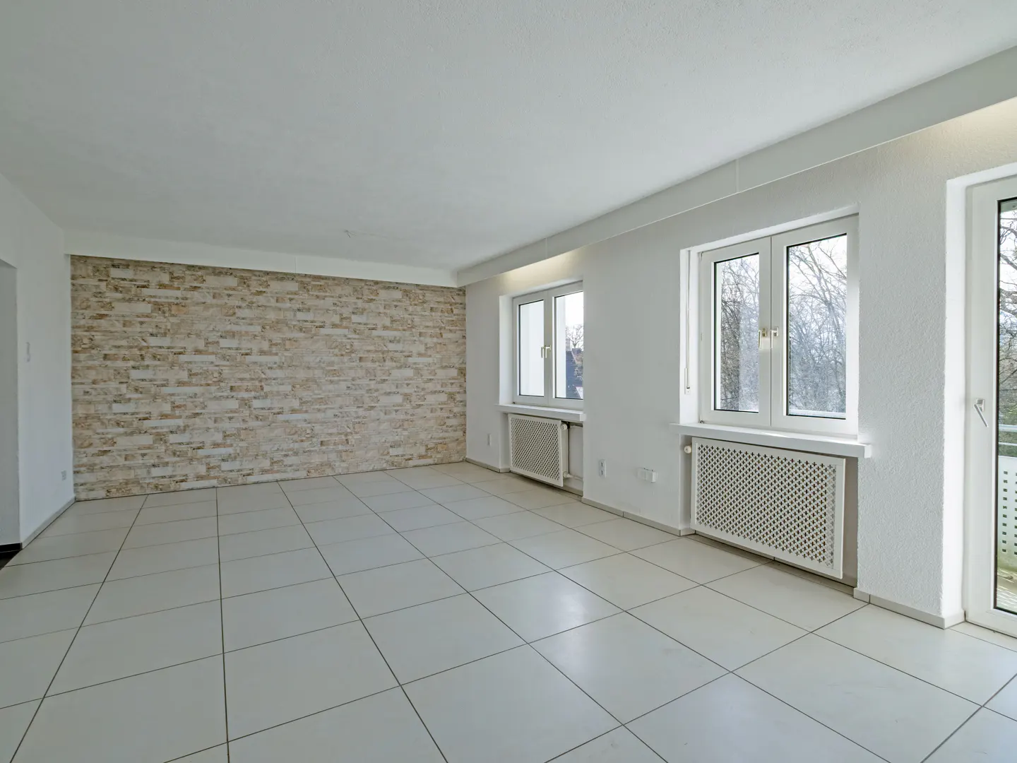 An empty, bright room with white tile floors, white walls, and a brick accent wall. Windows let in natural light.