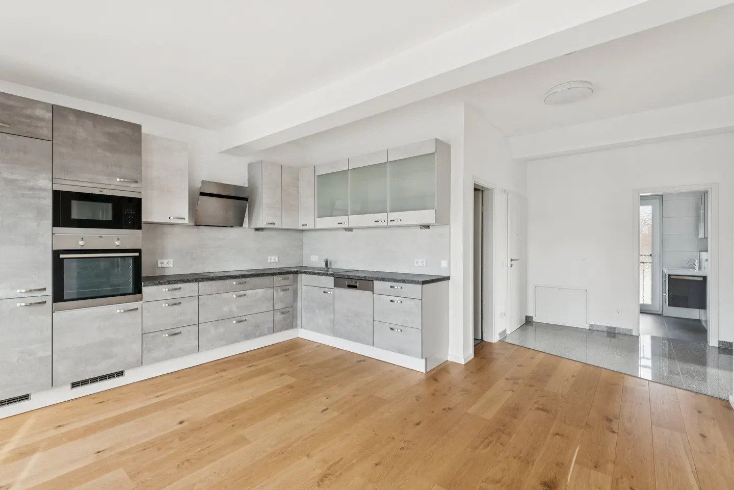 A bright, modern kitchen with gray cabinets, stainless steel appliances, and hardwood floors. A doorway leads to another room with gray tile.
