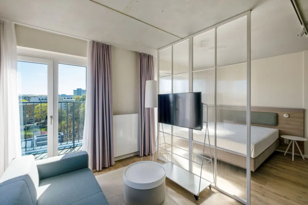 Bright studio apartment with a blue sofa, balcony, and a glass partition separating the living area from the bed.