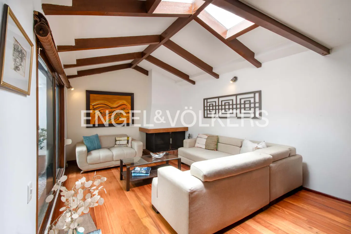 Living room with hardwood floors, white walls, and exposed beams. Two sofas face a fireplace. A skylight provides natural light.