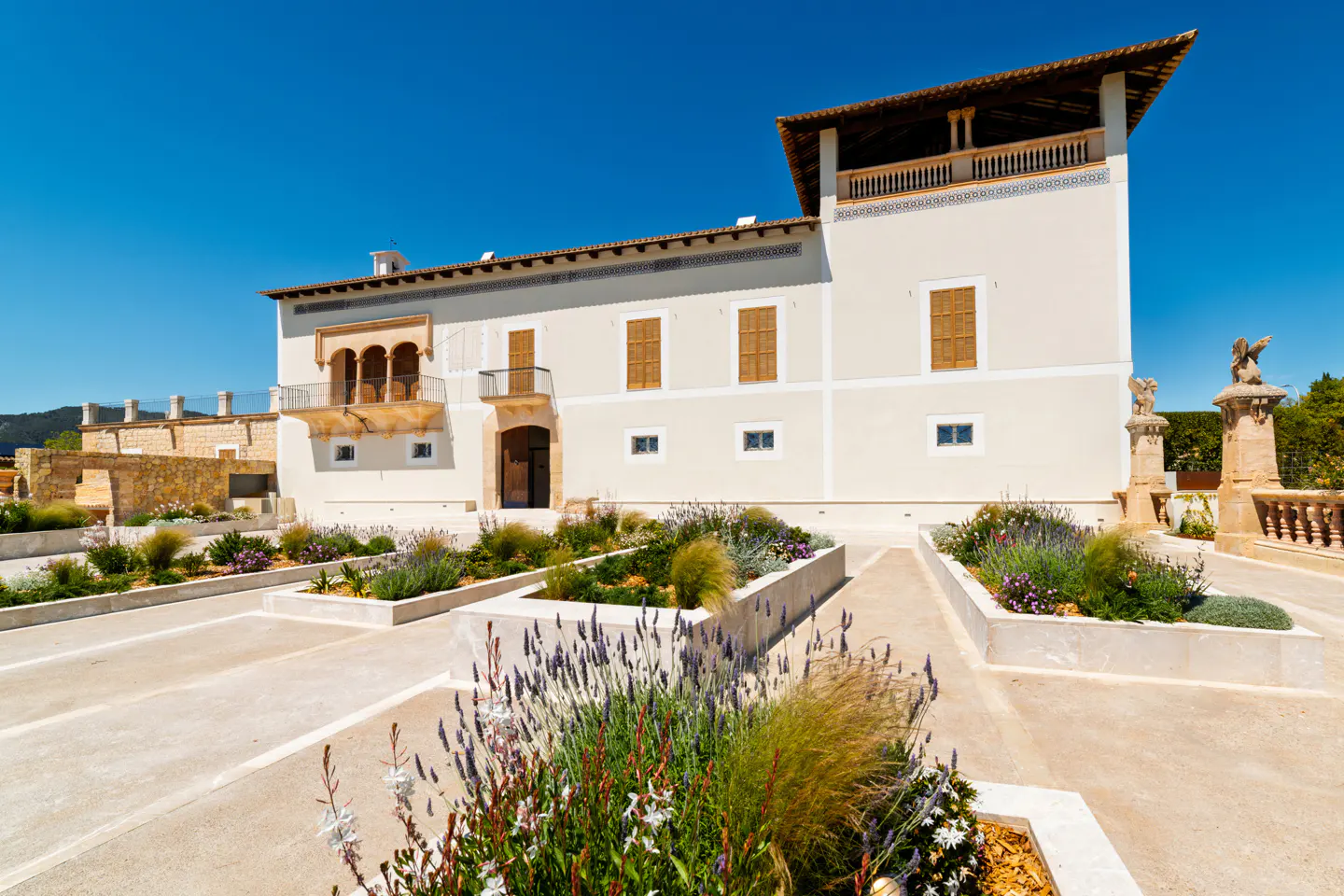 Exterior of a large, light beige house with brown shutters and a garden with lavender under a clear blue sky.