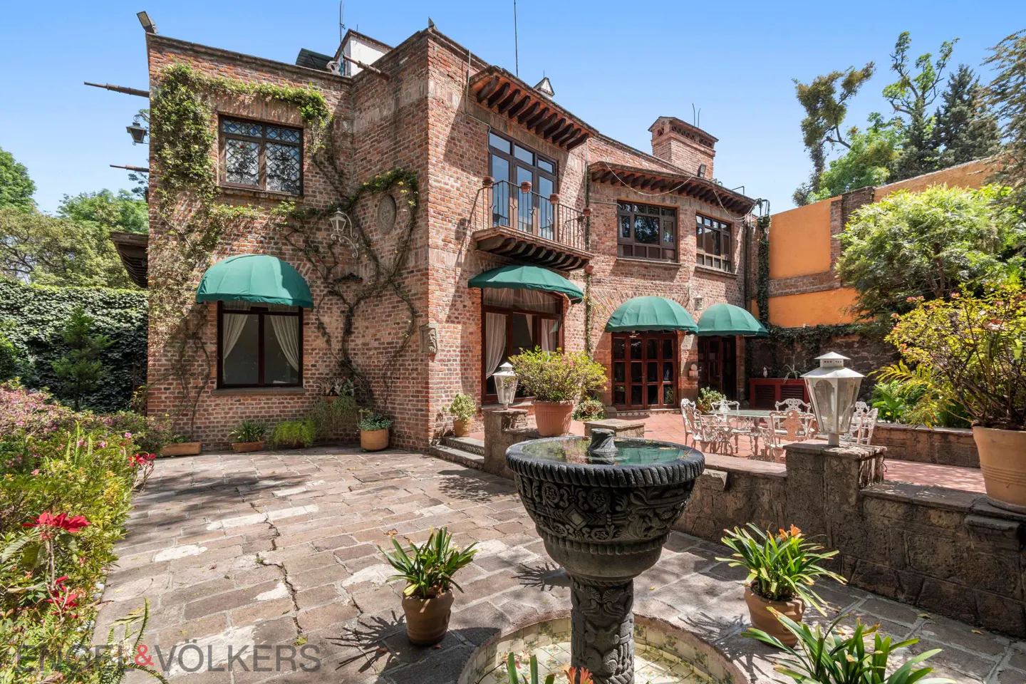 A rustic brick mansion with green awnings, a stone patio, a central fountain, and lush garden landscaping.