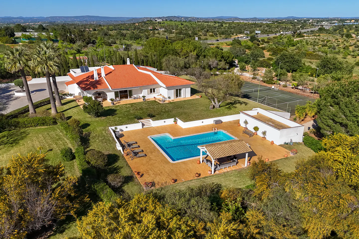 Aerial view of a white house with a red tile roof, pool, tennis court, and green landscape.