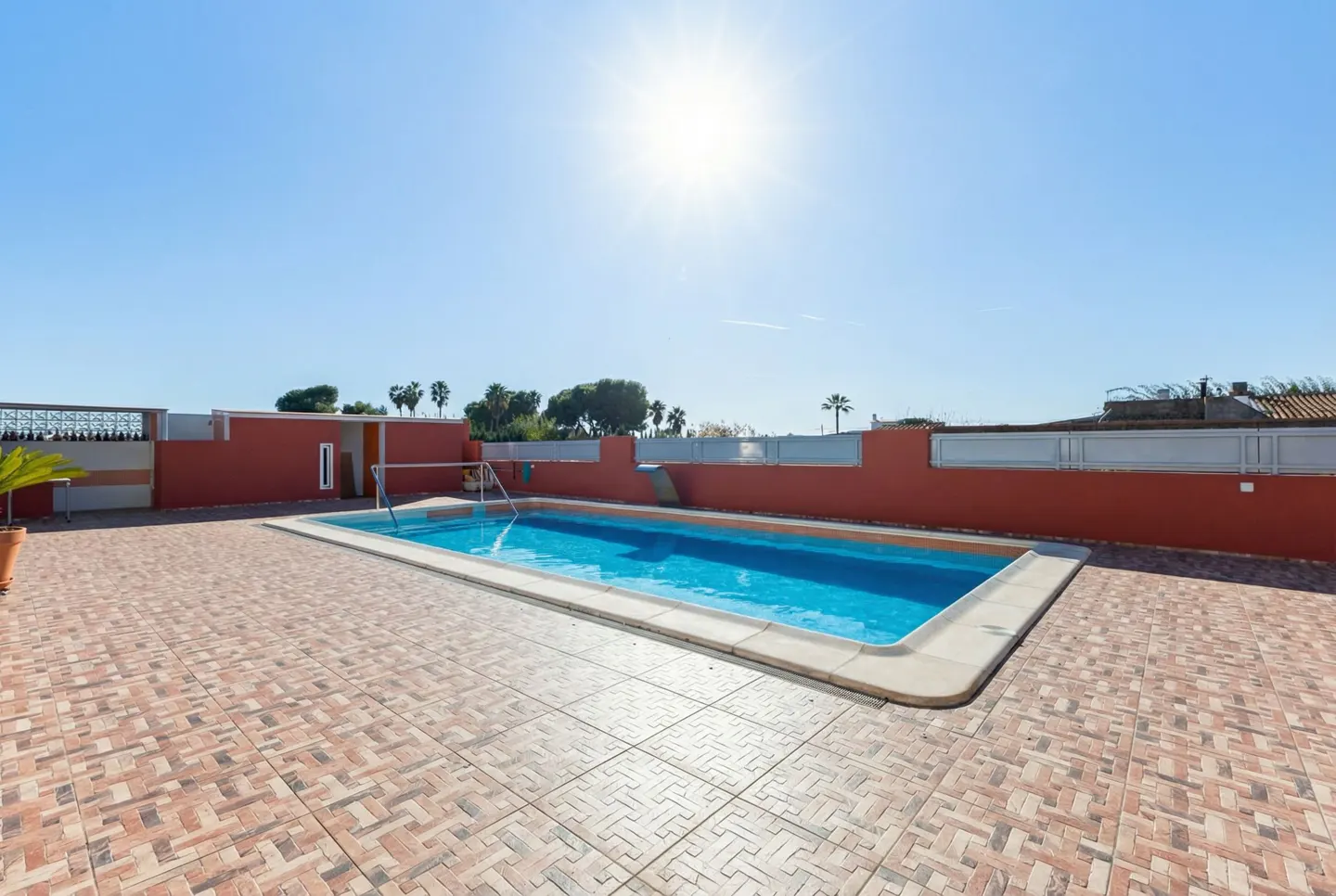 Outdoor pool with blue water, surrounded by a brick patio and a red wall under a clear blue sky.