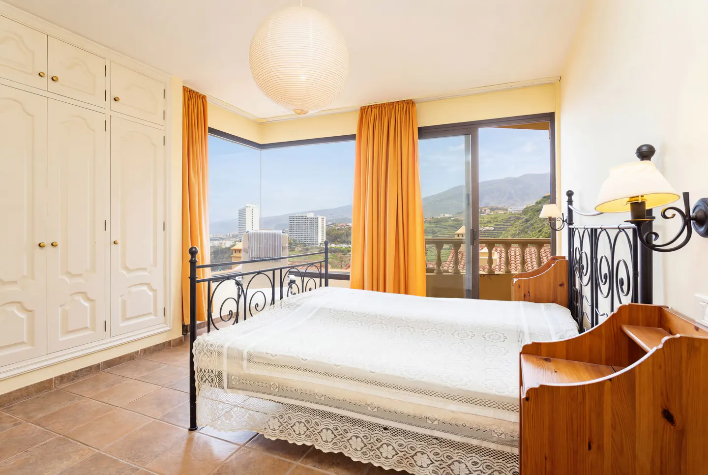 Bedroom with a black metal bed frame, white bedding, and orange curtains. A large window overlooks a city and mountains.