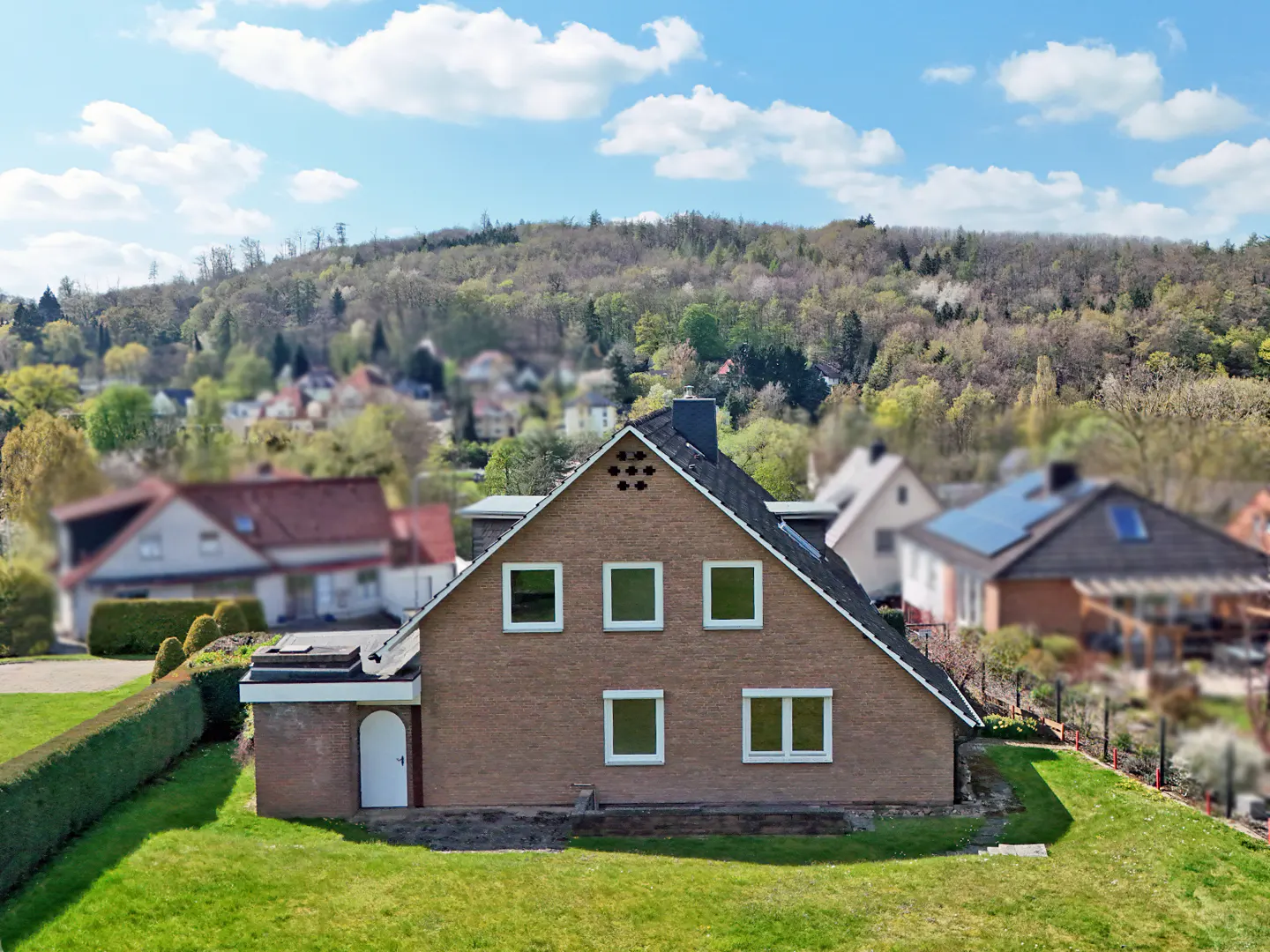 A tan brick house with a dark roof sits on a green lawn under a blue sky with white clouds.