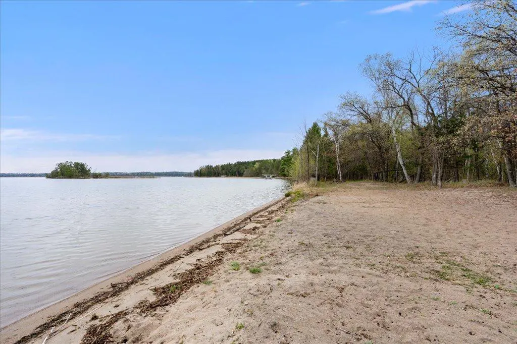Sandy beach meets calm lake under a blue sky. Trees line the shore, with a small island visible in the distance.