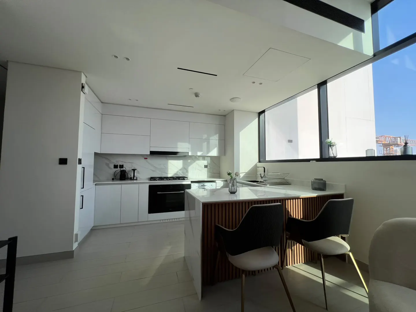 Modern white kitchen with marble backsplash, a wooden kitchen island with two chairs, and large windows with bright light.