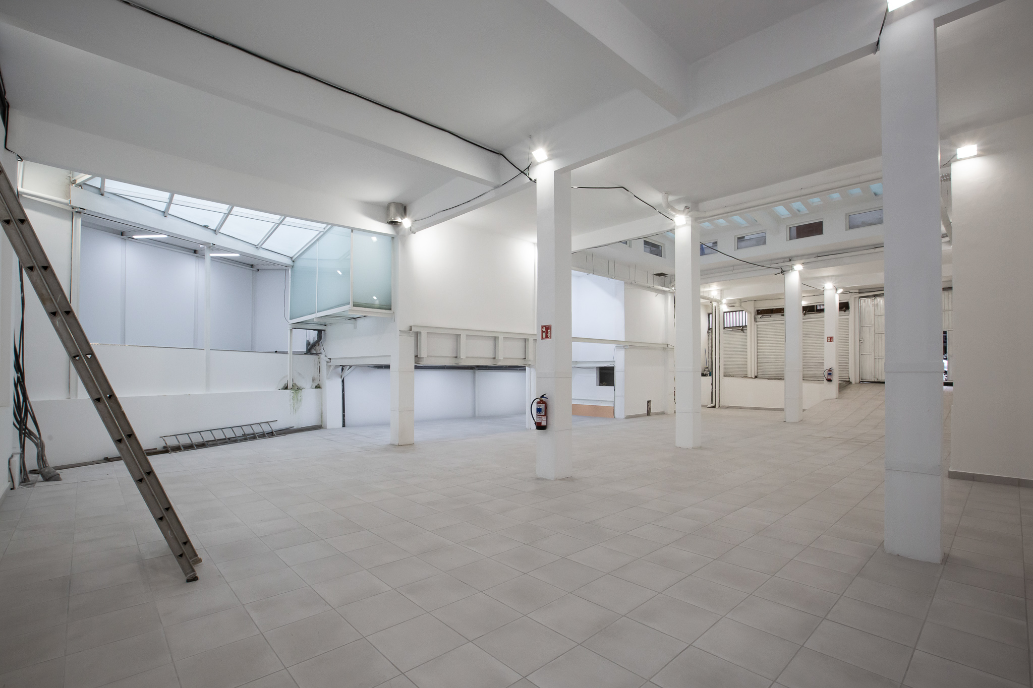 Bright, empty warehouse space with white walls, columns, and tile floor. A ladder leans against the wall. Skylights provide natural light.