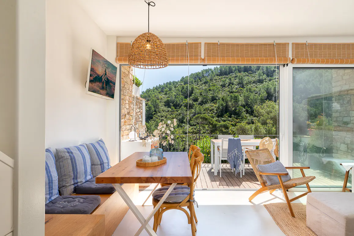 Bright dining area with a wooden table, blue striped cushions, and a woven pendant light. Large windows overlook a lush green hillside and outdoor seating.