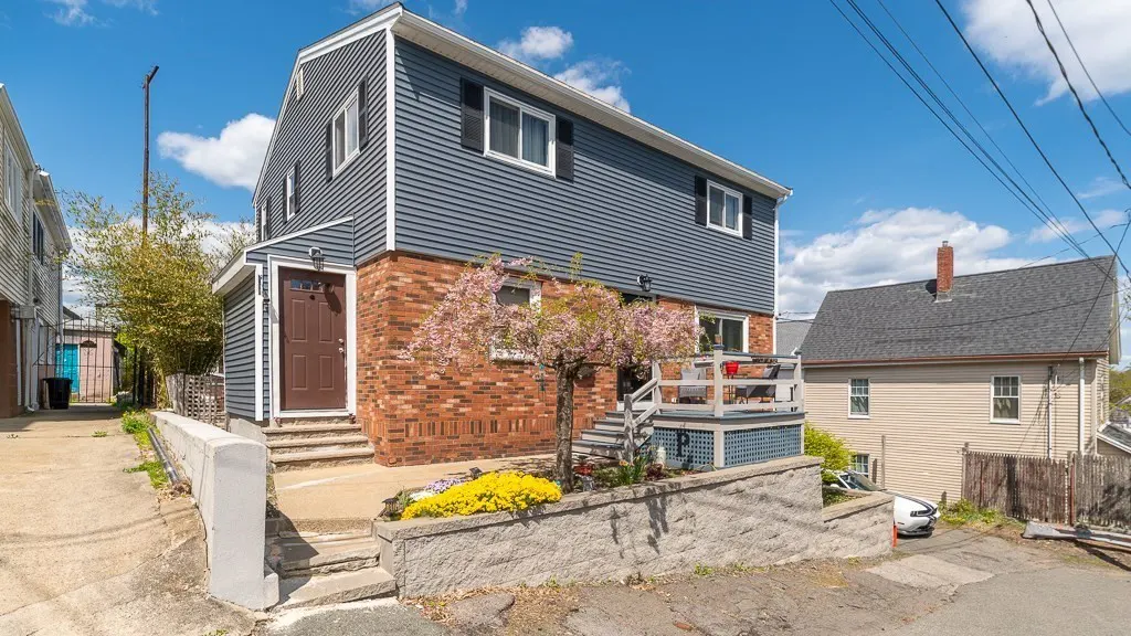 Two-story house with blue siding and brick facade, a small deck, and a brown front door. Yellow flowers in the front yard.