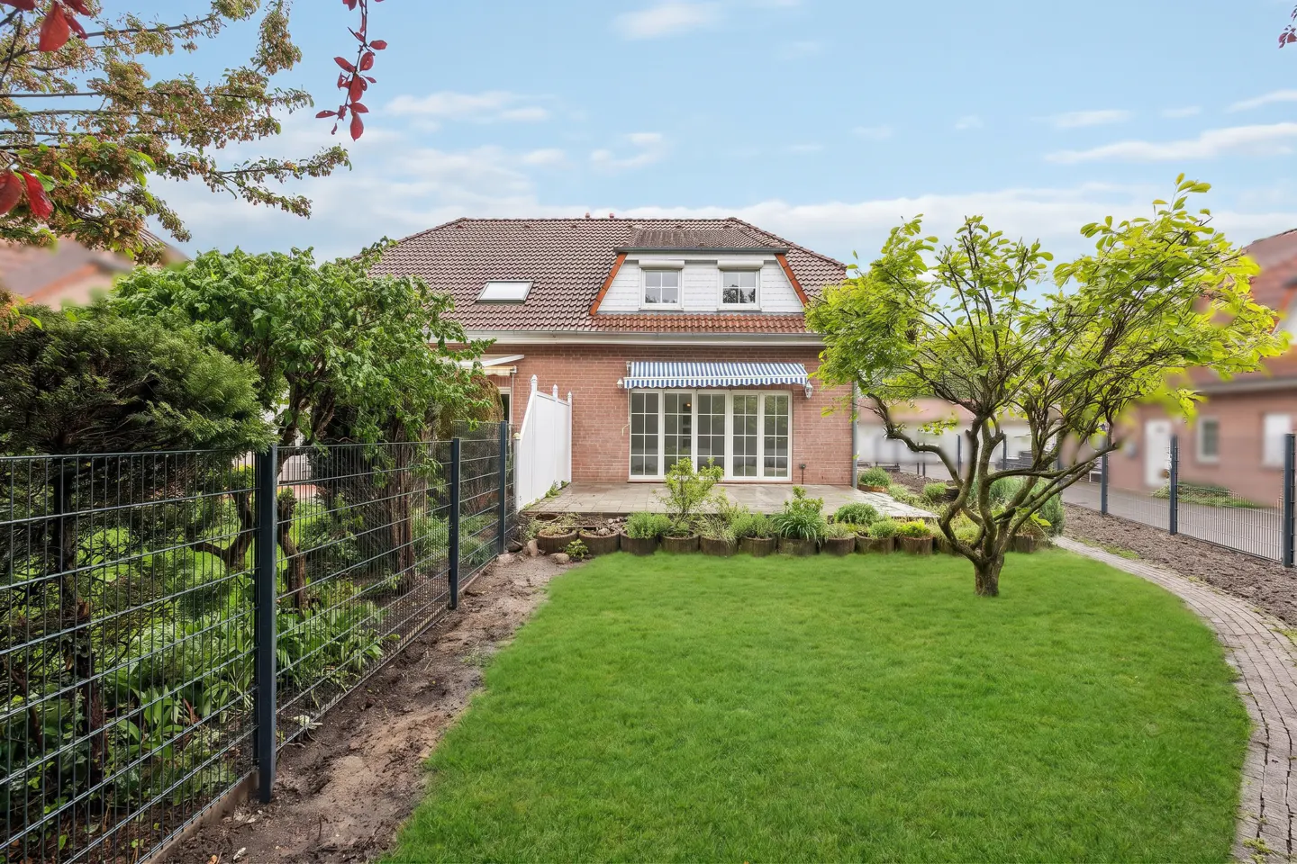 Backyard view of a brick house with a green lawn, trees, and a metal fence under a blue sky.