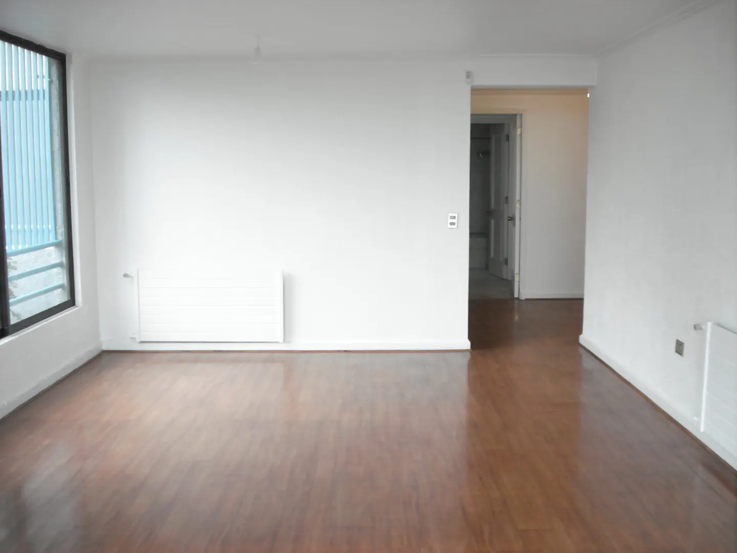 An empty room with white walls and dark wood floors. A window is on the left, and a doorway leads to a bathroom in the back.