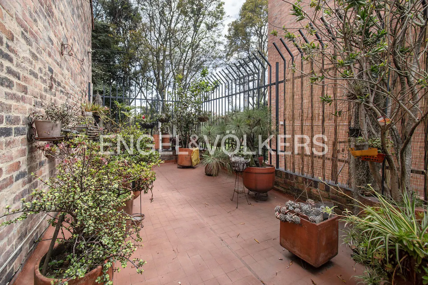 Outdoor patio with brick walls, terracotta tiles, and potted plants. A black metal fence separates the space from trees and sky.