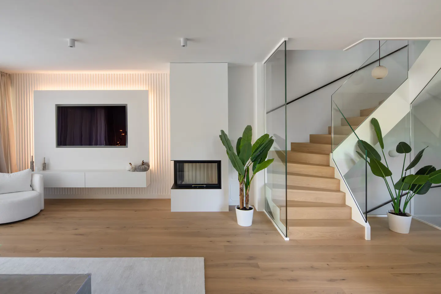 Bright living room with wood floors, a TV, fireplace, and stairs with glass railings. Potted plants add greenery.