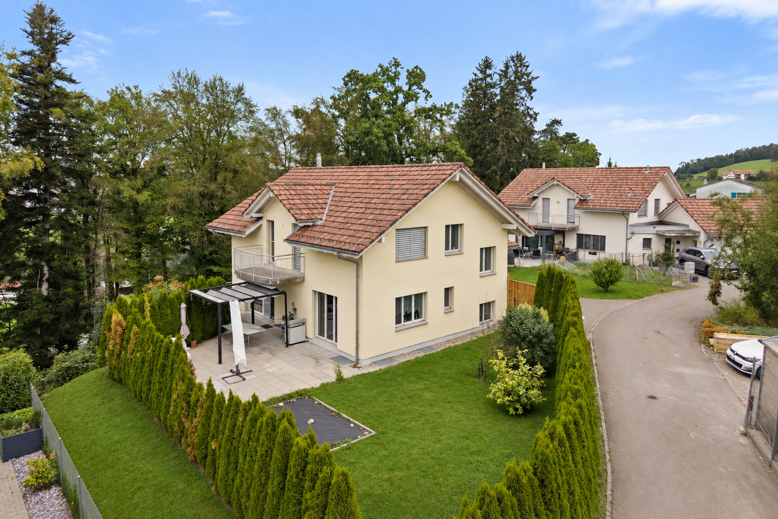 Two-story cream-colored house with a red tile roof, patio, and green lawn enclosed by tall hedges on a sunny day. [AI-generated text]