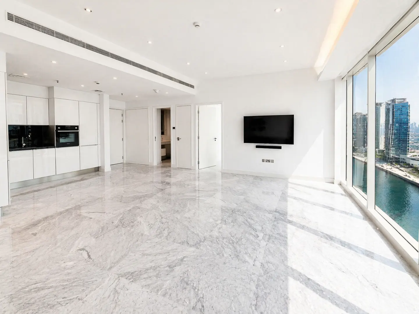 Bright, modern apartment with marble floors, white walls, and a large window overlooking a city skyline and waterway. A flat-screen TV is mounted on the wall.
