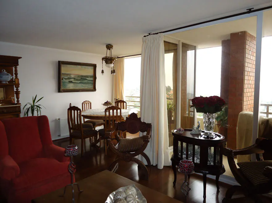 A bright living room with a red armchair, wooden furniture, and a painting of the ocean on the wall. A balcony is visible through a sliding glass door.