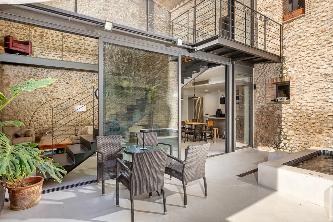 Stone house patio with table, chairs, and jacuzzi. Interior view through glass doors shows stairs, kitchen, and living area.