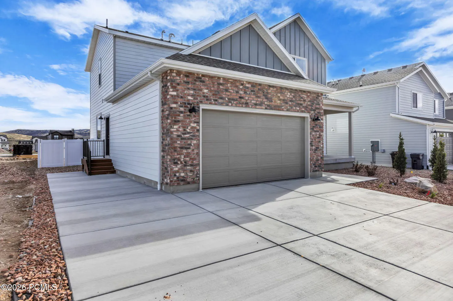 Two-story house with a gray garage door and brick facade. A concrete driveway leads to the garage. Blue sky with clouds.