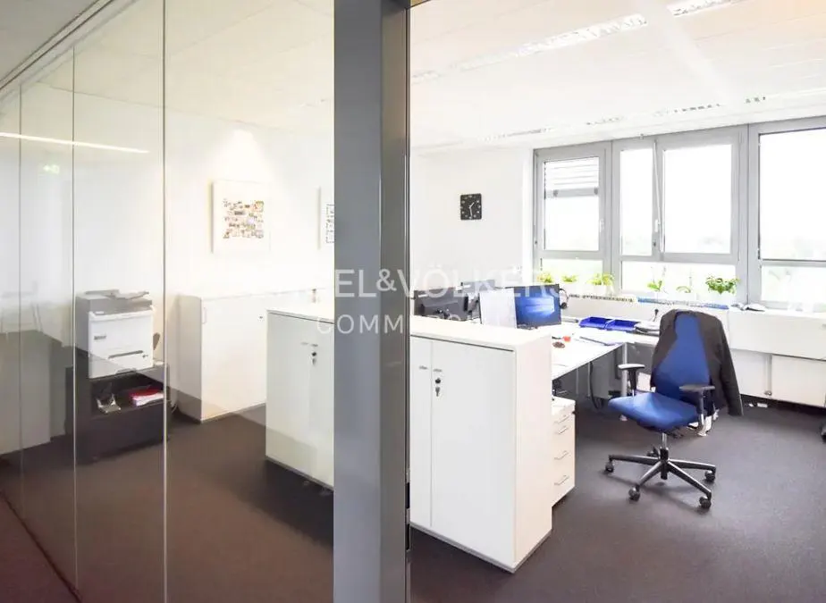 Bright office space with white walls, black carpet, and large windows. Desks, cabinets, and a blue chair are visible.