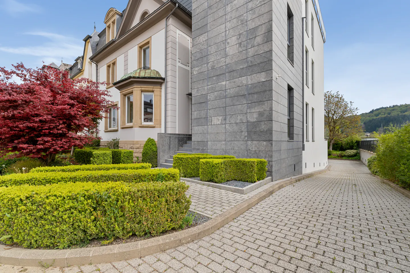 Exterior of a modern home with gray stone and white siding, green hedges, and a red tree. A brick driveway leads to the house.