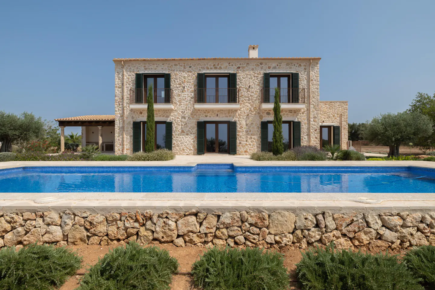 Two-story stone house with dark shutters overlooks a blue swimming pool. A stone wall borders the pool, with green bushes in front.