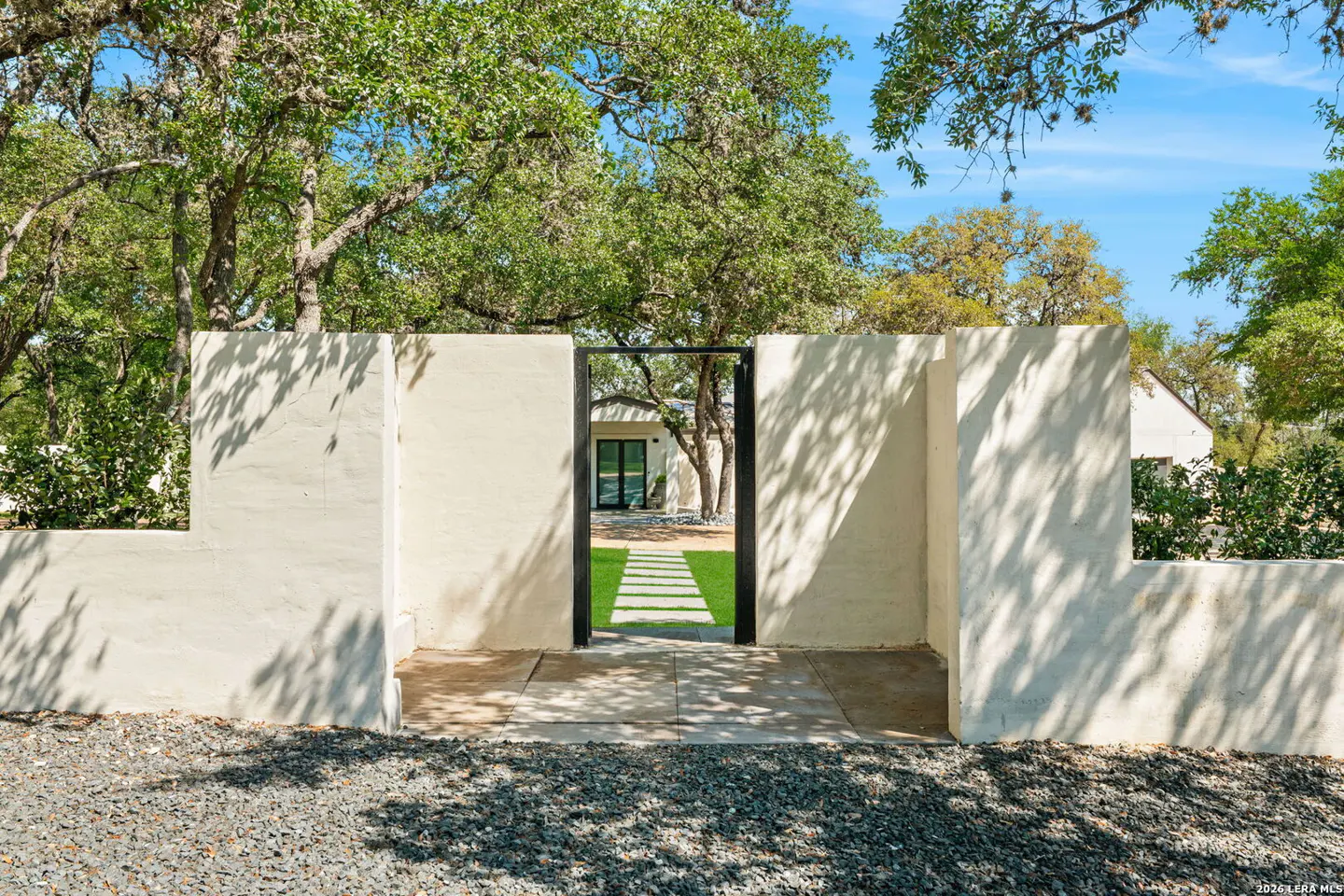 Exterior view of a home's entrance with a white wall and black metal gate. A stone path leads to the house, surrounded by green grass and trees.