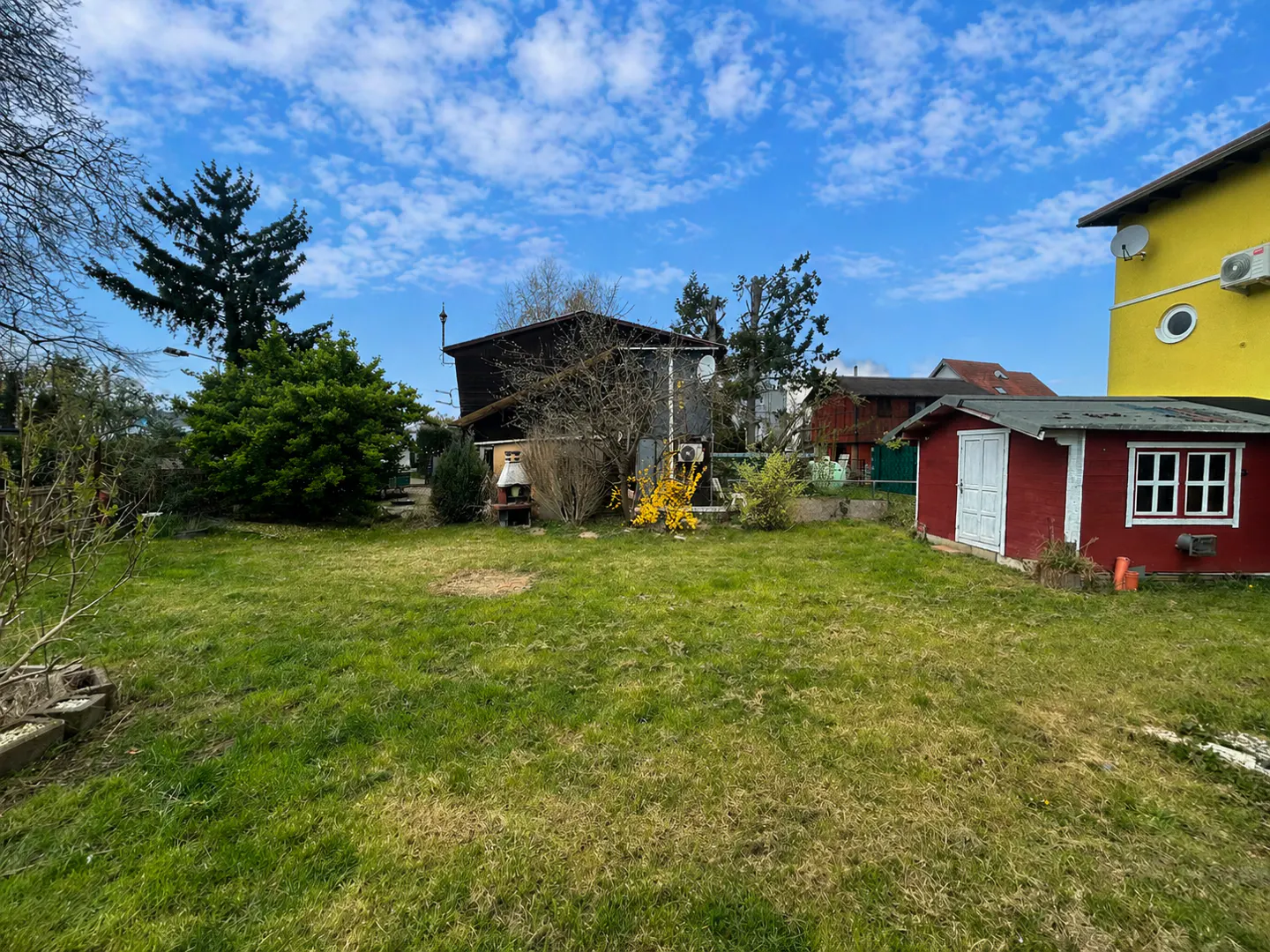 A green lawn with a red shed and yellow house under a blue, cloudy sky.