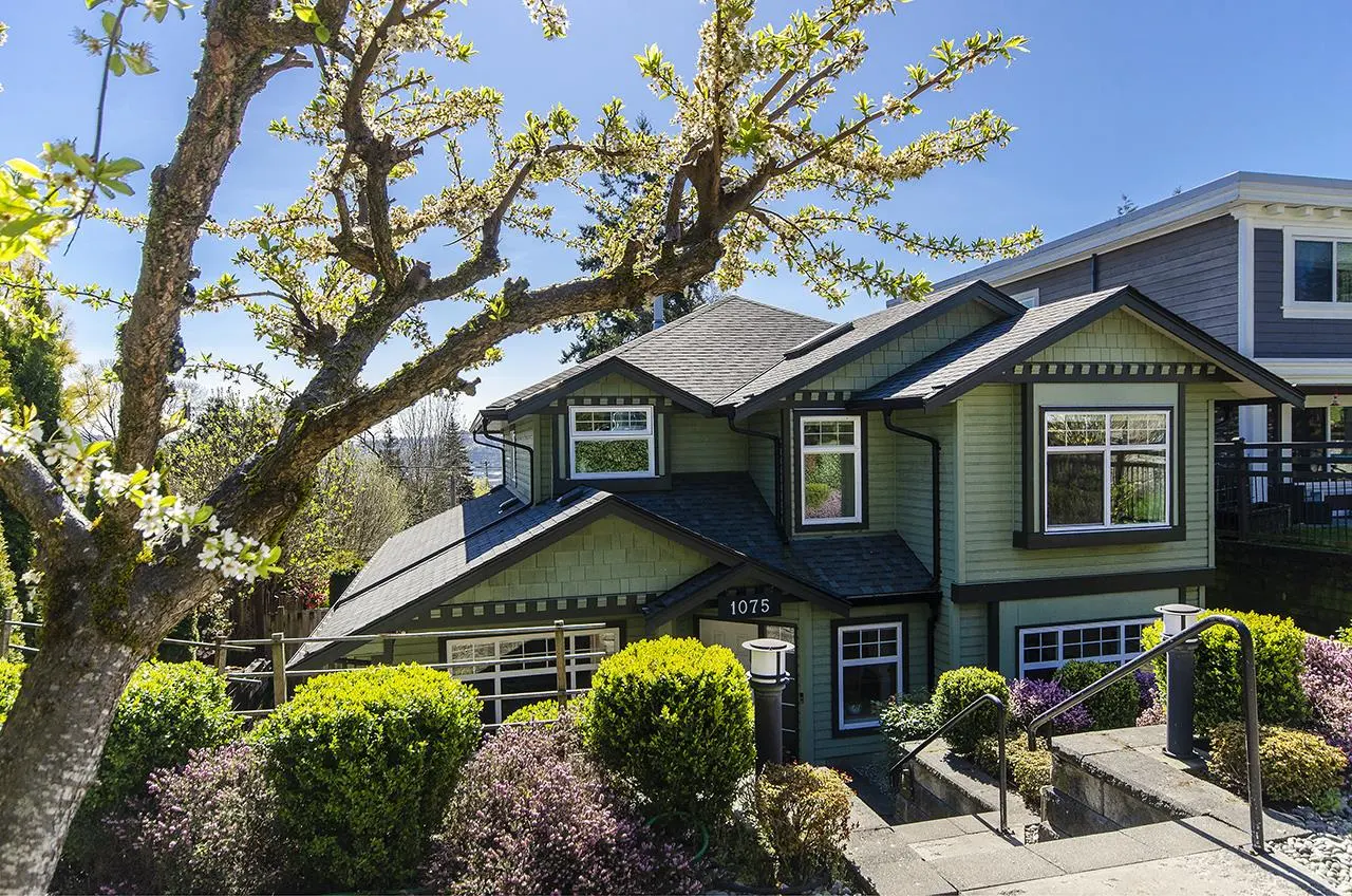 Two-story green house with black roof and white trim, number 1075, surrounded by bushes and a flowering tree.