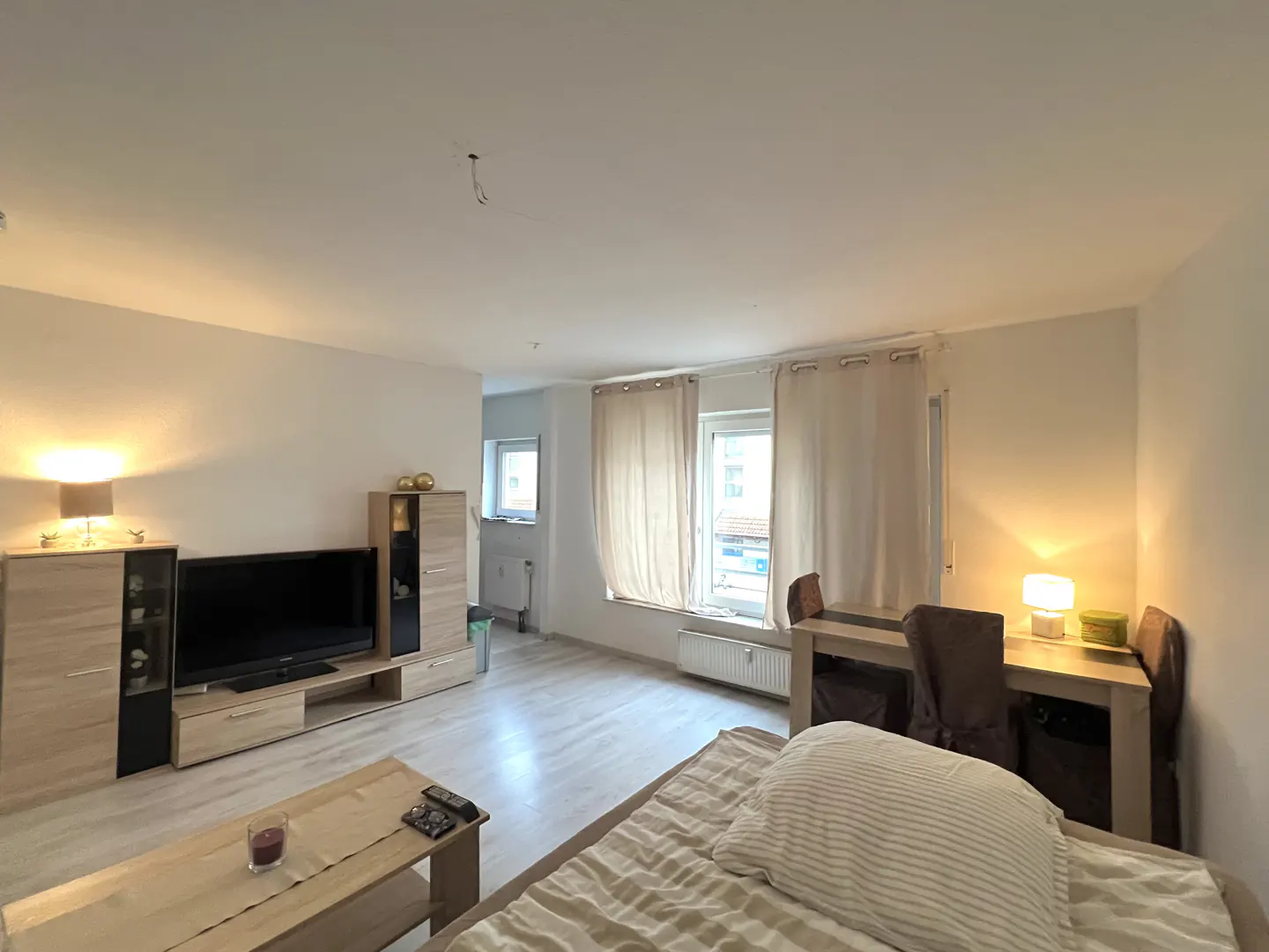 Bright living room with light wood floors, a TV unit, a coffee table, and a dining area with beige curtains.