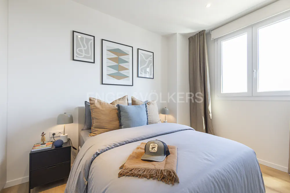 Bedroom with a bed, blue duvet, tan throw, and a baseball cap. Three framed art prints hang above the bed. A black nightstand is on the left.