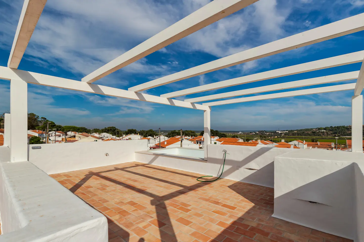 Rooftop patio with brick flooring, white pergola, and a small pool. A green hose is coiled near the pool. Houses with orange roofs are in the background.