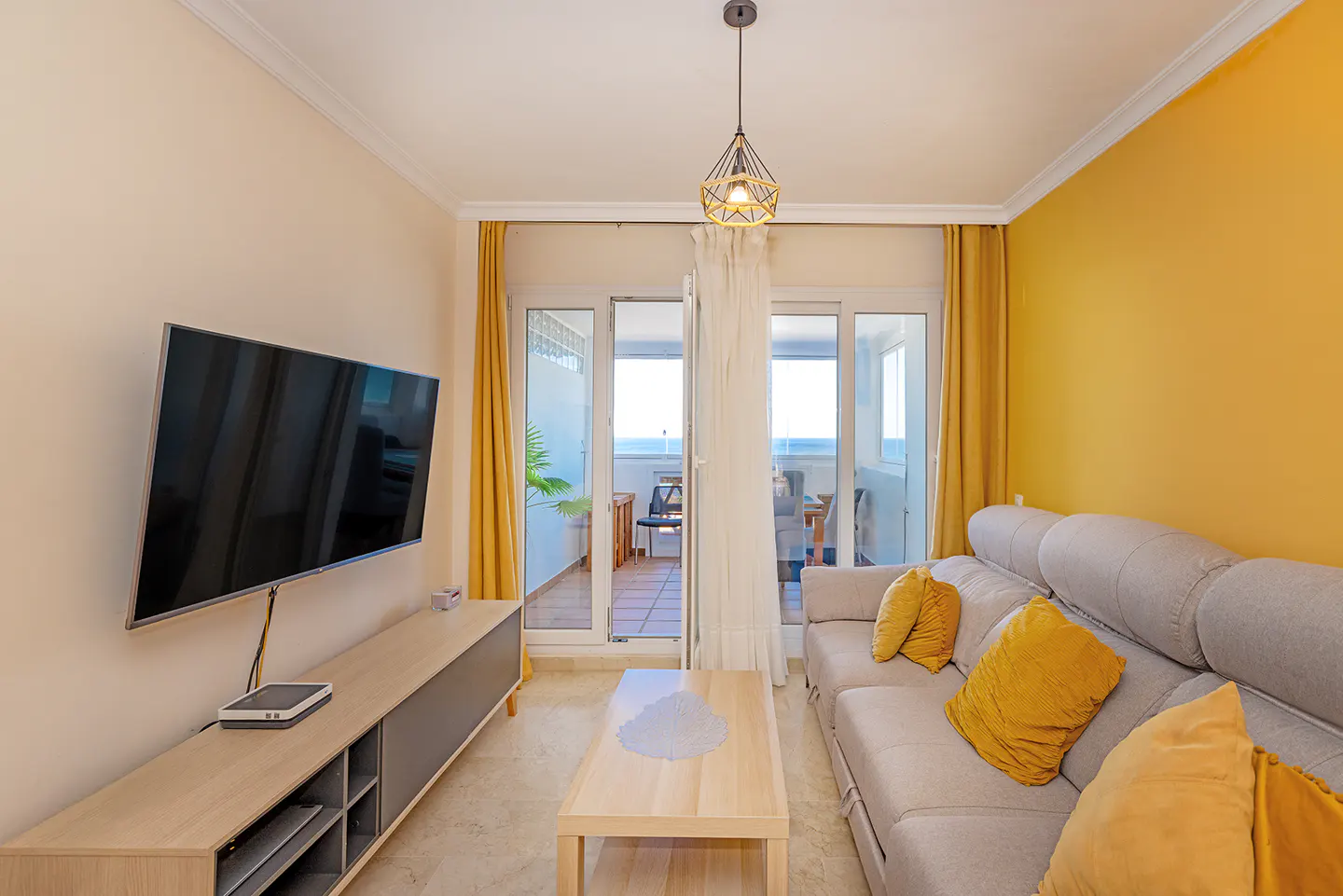 Bright living room with a gray sofa, yellow pillows, and a TV. Balcony doors open to an ocean view.