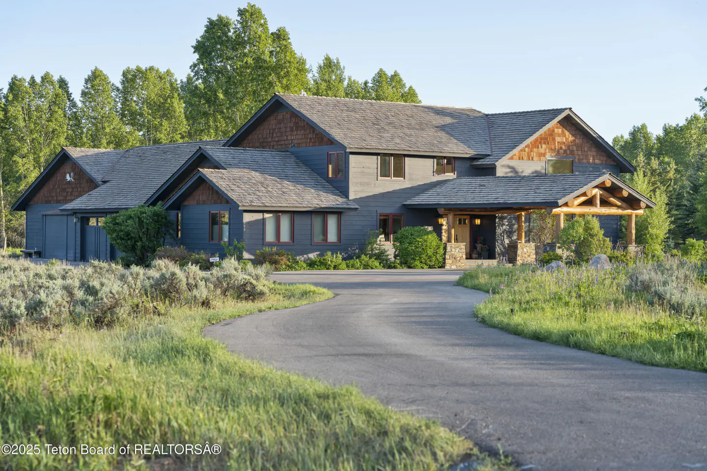 Exterior view of a large, two-story gray house with a log-beamed porch and a winding driveway. Trees are in the background.