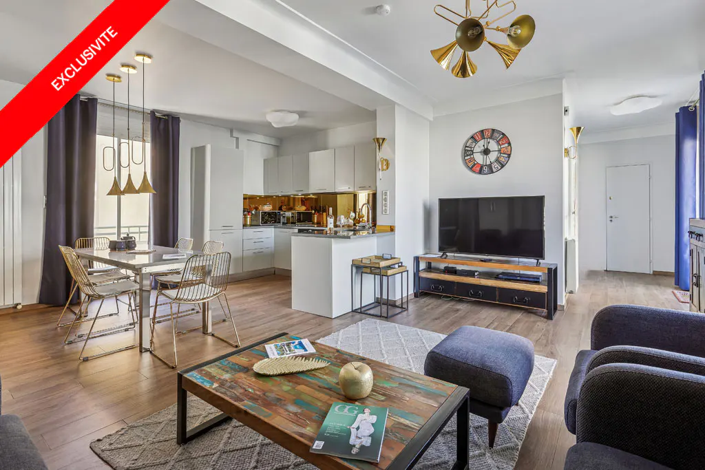 Open-concept apartment with a dining table, white kitchen, TV, and living area with a colorful coffee table. "Exclusivite" banner.