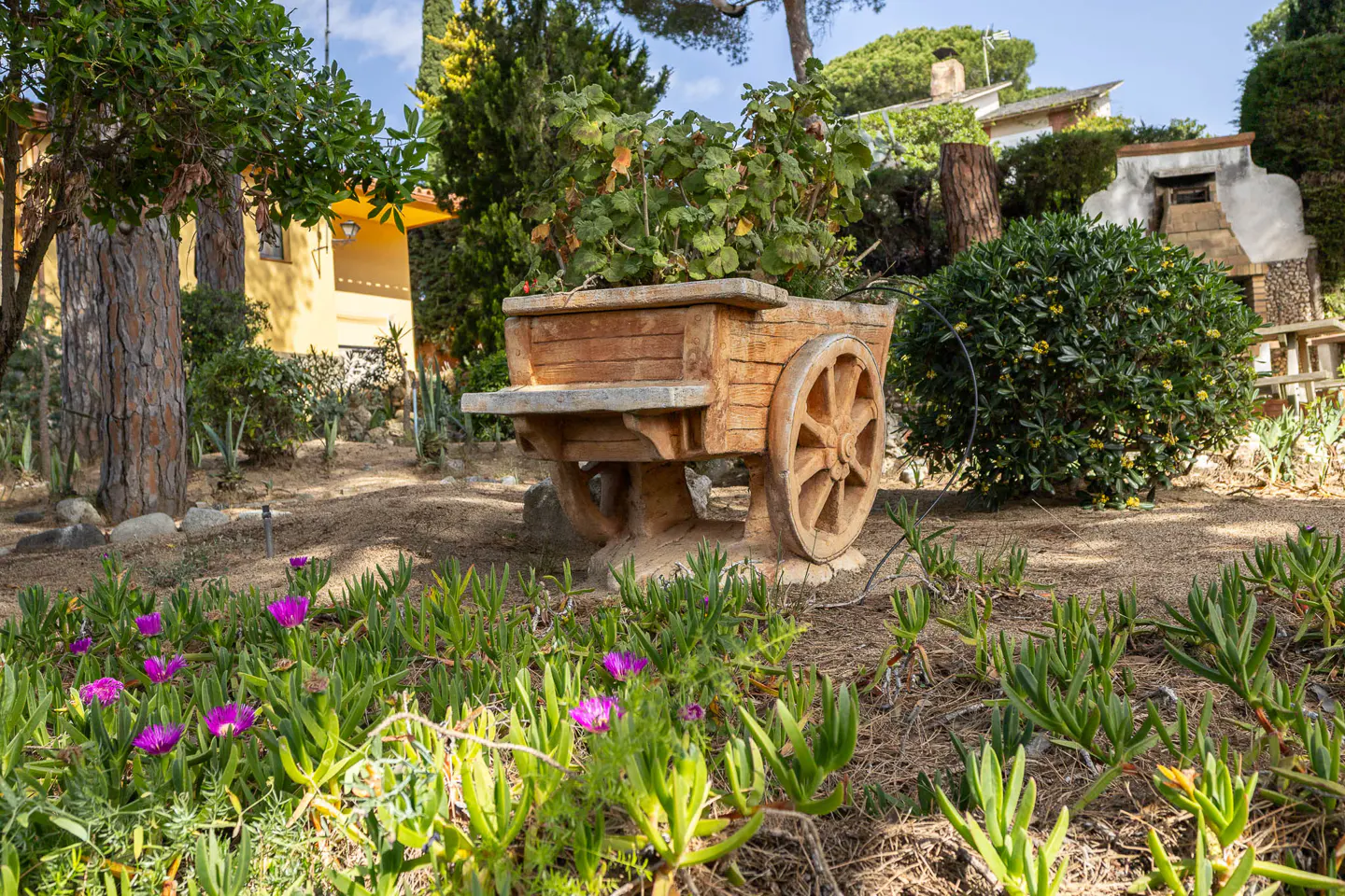 A wooden wagon planter filled with greenery sits in a garden with purple flowers and a yellow house in the background.